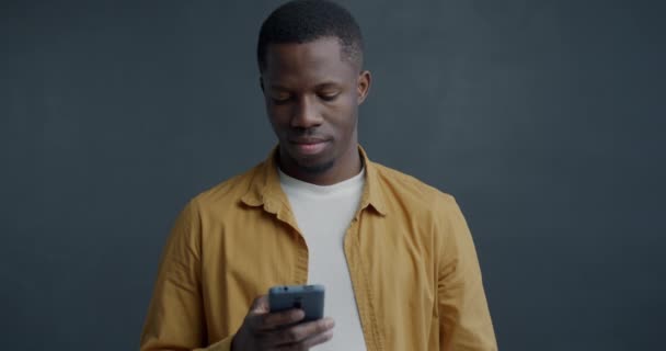 グレーの背景にソーシャルメディアを使用してスマートフォンメッセージを使用するアフリカ系アメリカ人のゆっくりとした動きの肖像画 人とガジェットのコンセプト — ストック動画