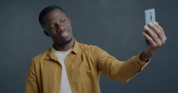 グレー色の背景で楽しんでいるスマートフォンカメラのためにポーズを取る遊び心のあるアフリカ系アメリカ人の男のゆっくりとした動きの肖像画 — ストック動画