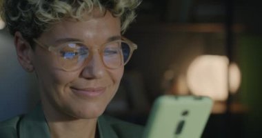 Gecenin karanlığında akıllı telefon kullanarak internette sohbet eden gülümseyen genç iş kadınının yakın çekimi. Gadget ve işadamları kavramı.