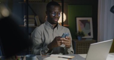 Afrika kökenli Amerikalı iş adamı akıllı telefonuyla mesaj atıyor ve akşamları karanlık ofisteki masasında alet kullanarak gülümsüyor. İnsanlar ve internet bağlantısı kavramı.