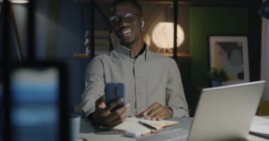 Neşeli Afro-Amerikan iş adamı akıllı telefon ve kulaklıkla video görüşmesi yapıyor ve akşamları karanlık ofiste gülüyor.