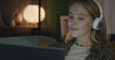 Kablosuz kulaklıkla müzik dinleyen ve geceleri karanlık oturma odasında dizüstü bilgisayarla çalışan genç bir bayan. Modern teknoloji ve milenyum kavramı.