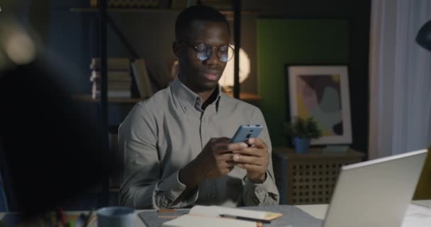 アフリカ系アメリカ人のビジネスマンは 夜の暗いオフィスの机でスマートフォンと笑顔でテキストを送ります 人とインターネットのコンセプト — ストック動画