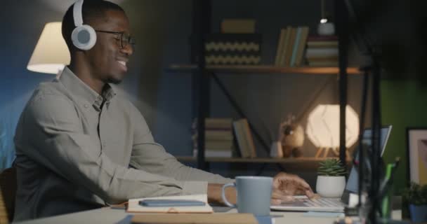 アフリカ系アメリカ人男性がヘッドフォンを着て音楽を聴き 夜遅くに働くダークオフィスでラップトップを使用しています ビジネスと職場の活動コンセプト — ストック動画