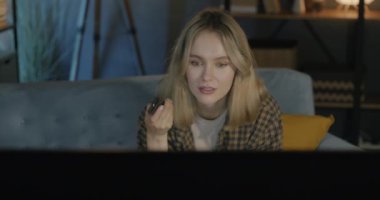 Neşeli genç bir kadın TV izlerken gülümsüyor ve geceleri karanlık bir dairede kanepede oturuyor. Halk ve kitle iletişim araçları.