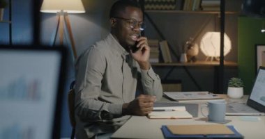 Neşeli Afrikalı Amerikalı çalışan cep telefonuyla konuşuyor. Geceleri ofisteki masada gülüyor ve kahve içiyor. İletişim ve iş konsepti.