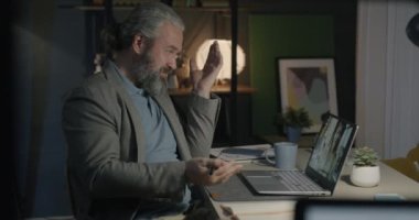 Neşeli, olgun iş adamı bilgisayarla video görüşmesi yapıyor ve geceleri karanlık ofiste bayan meslektaşlarıyla konuşuyor. İletişim ve iş konsepti.