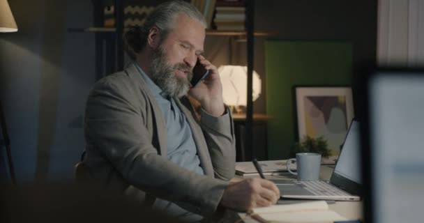 ビジネスマンは 携帯電話を作って 夜にオフィスでノートブックにノートを書く ビジネスコミュニケーションとナイトシフトのジョブコンセプト — ストック動画