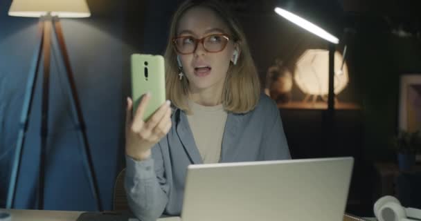 スマートフォンやイヤホンを使ったオンラインビデオ通話を夜間オフィスで行うキャリーフの若いビジネスマン ビジネスディスカッションと現代技術コンセプト — ストック動画