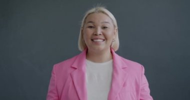 Umut dolu Asyalı bir kadının gri arka plandaki kameraya bakarak dilek tutmasını gösteren yavaş çekim portresi. Başarılı kişi ve olumlu duygu konsepti.