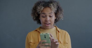 Akıllı telefon kullanan heyecanlı Afrikalı Amerikalı kadının yavaş çekimde portresi. Gri arka planda iyi haberlerin tadını çıkarıyor.