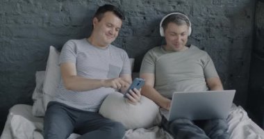 Pijamalı neşeli eşcinsel çift dizüstü bilgisayar kullanıyor kulaklıklı müzik dinliyor ve evde akıllı telefonlu sosyal medyanın tadını çıkarıyor. Modern teknoloji ve homoseksüel ilişki kavramı.