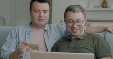 Neşeli eşcinseller internet mağazasında internet kartı alışverişi yaparak online ödeme yapıyorlar ve evde gülüyorlar. LGBTQ ilişkisi ve tüketim kavramı.