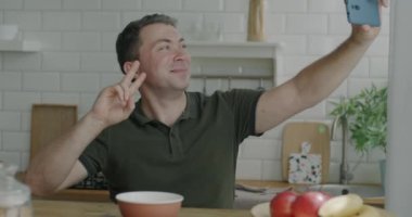 Neşeli adam akıllı telefon kamerasıyla selfie çekiyor. Mutfakta baş parmak hareketleriyle poz veriyor. Modern teknoloji ve fotoğraf konsepti.