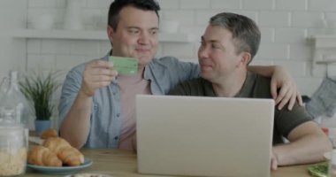 İnternetten alışveriş yapan mutlu eşcinsel çift dizüstü bilgisayar ve kredi kartı konuşmalarıyla mutfaktaki mutluluğu ifade ediyor. Bankacılık ve aynı cinsiyet ilişkisi.