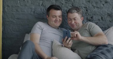 Neşeli eşcinsel çift yatakta uzanıp akıllı telefon kullanıyor ve boş zamanlarında sosyal medya içeriğinin keyfini çıkarıyorlar. Gadget ve homoseksüel ilişki kavramı.