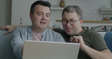 Neşeli eşcinsel çift dizüstü bilgisayar kullanarak evdeki kanepede oturup konuşuyorlar. Modern teknoloji ve çevrimiçi içerik kavramı.