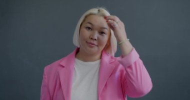 Saçlarıyla oynayan ve gri arka planda kameraya bakan sevimli Asyalı kadının yavaş çekim portresi. İnsanlar ve olumlu duygu kavramı.