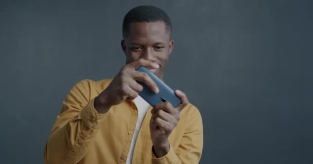 グレーの背景で楽しんでいるスマートフォンでビデオゲームをしているアフリカ系アメリカ人のゆっくりとした動きの肖像画 ゲームとエンターテイメントのコンセプト — ストック動画