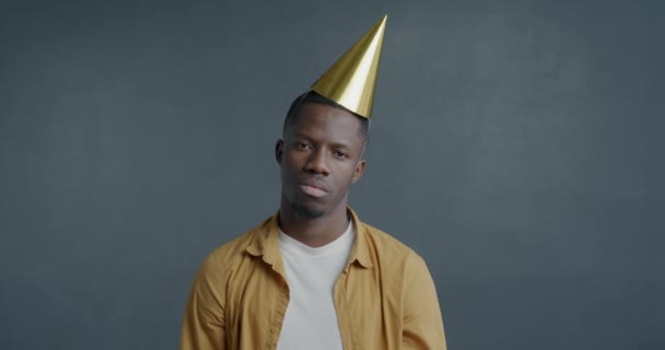 不幸なアフリカ系アメリカ人の男性のゆっくりとした動きの肖像画は パーティーの帽子を脱ぎ 灰色の背景に退屈な気分を吹く — ストック動画