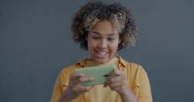Neşeli Afro-Amerikan kadın portresi çevrimiçi video oyunu oynayan akıllı telefonlu gri arka planda eğlenen. Gadget ve eğlence konsepti.