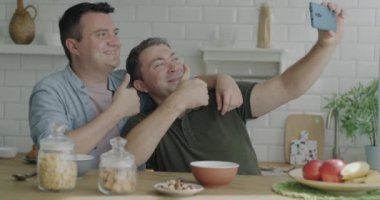 Neşeli eşcinsel çift akıllı telefon kameralı selfie çekiyorlar. Evde mutfaklarında baş parmakları havada poz veriyorlar. Erkekler fotoğraf çekiyor, sarılırken eğleniyor..
