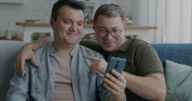 Akıllı telefondan gülümseyen eşcinseller fotoğraf izleyip evdeki koltukta el hareketi yapıyorlar. Modern aletler ve aynı cinsiyet ilişkisi kavramı..