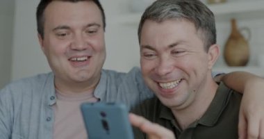 Akıllı telefon kullanan neşeli eşcinseller ekranlara bakıp gülüyor ve evde online içeriğin keyfini çıkarıyorlar. Modern cihazlar ve LGBTQ ilişki konsepti.