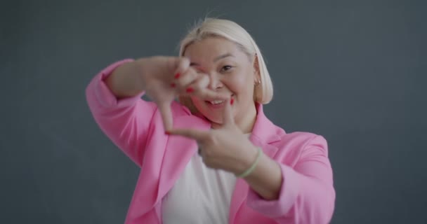 灰色の背景に正方形の手のジェスチャーを作る想像上のビューファインダーを通してカメラを見ている創造的なアジアの女性のゆっくりとした動きの肖像画 — ストック動画