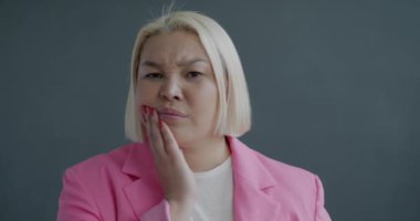 Diş ağrısı çeken, yanağa dokunan, çatık kaşlı, gri arka planda kameraya bakan mutsuz Asyalı kadının ağır çekimde portresi.