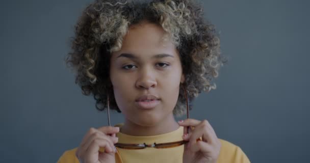 遊び心のあるアフリカ系アメリカ人女性のクローズアップスローモーション肖像画は スタイリッシュなサングラスを着て 灰色の裏地でカメラを眺める — ストック動画