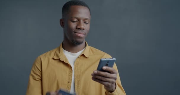 グレーの背景にスマートフォンとクレジットカードでオンラインショッピング幸せなアフリカ系アメリカ人男性のゆっくりとした動きの肖像画 ビジネスとモダンなライフスタイルコンセプト — ストック動画
