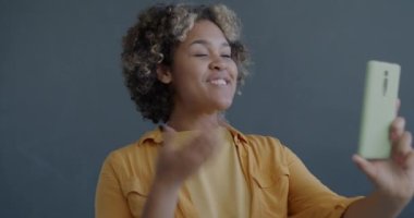 Neşeli Afrikalı Amerikalı kadın el sallayarak gri arka planda akıllı telefonuyla çevrimiçi video görüşmesi yapıyor. İnsanlar ve iletişim konsepti.