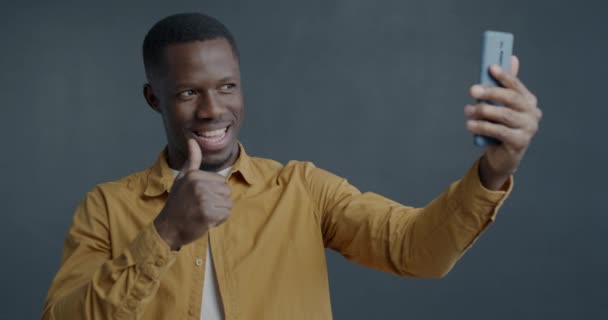 幸せなアフリカ系アメリカ人の男の肖像画 親指を示すスマートフォンでオンラインビデオ通話を行い グレー色の背景で話すOkハンドジェスチャー — ストック動画