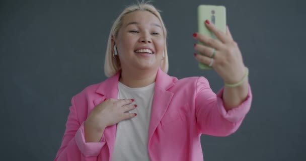スマートフォンとワイヤレスイヤホンを使用してオンラインビデオ通話をする陽気なアジアの女性のゆっくりとした動きの肖像画は 灰色の背景で笑います — ストック動画