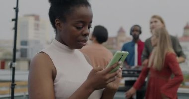 Neşeli Afrikalı Amerikalı kadın akıllı telefon kullanıyor ve çatıdaki partide arkadaşlarıyla gülümsüyor. İnsanlar farklı gruplarda dans edip arka planda eğleniyorlar..