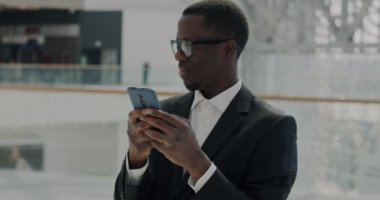 Neşeli bir Afro-Amerikan iş adamı modern cam duvar ofis merkezinde akıllı telefon kullanarak mesajları kontrol ediyor. İletişim ve teknoloji kavramı.
