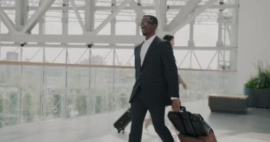 Zarif Afro-Amerikan iş adamının çantalı insanlar etrafta dolaşırken havaalanında çantasıyla dolaşması. Modern şehir panoramik pencerelerde görünür.