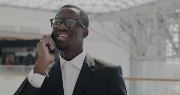 笑着穿西装的非洲裔美国商人在手机上讨论在阳光照射下的办公楼大堂进行合作 沟通和现代员工概念 — 图库视频影像