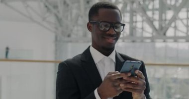 Takım elbiseli başarılı Afro-Amerikan çalışanın portresi ofis merkezinde panoramik pencereli akıllı telefonuyla geziyor. İş adamı ve teknoloji kavramı.