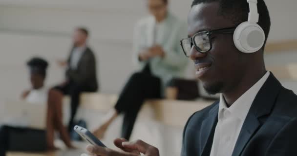 非裔美国商人通过耳机听音乐 并使用智能手机在办公楼附近的室外享受社交媒体 技术和工间休息概念 — 图库视频影像