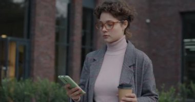 Neşeli genç bayan sokakta akıllı telefon kullanarak yürüyor internette sohbet ediyor ve kahve içmek için içiyor. Modern yaşam tarzı ve aletler kavramı.