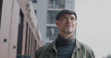 Şehir caddesinde tek başına duran şehir binalarını izleyen dalgın genç adam yavaş hareket ediyor. Modern metropol ve pozitif duygu kavramı.