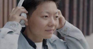 Şehir sokağında müzik dinlerken kulaklık takan neşeli Asyalı adamın yavaş çekim portresi. Modern teknoloji ve gençlik kültürü kavramı.