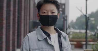 Sokakta salgın sırasında kameraya bakan yüz maskesi takan Asyalı adamın yavaş çekim portresi. Sağlık ve kentsel yaşam tarzı kavramı.