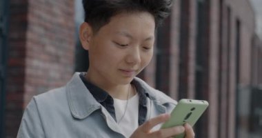Sosyal medyadan keyif alan kaygısız Asyalı öğrenci ekranlara dokunuyor ve şehrin dışında gülümsüyor. Modern aygıt ve çevrimiçi iletişim konsepti.