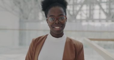 Afro-Amerikalı neşeli iş kadınının yavaş çekimde portresi. Geniş ofis binasında durmuş gülümsüyor. Cam duvarları var ve başarılı bir kariyerin tadını çıkarıyor.