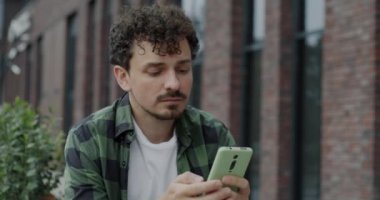 Akıllı telefon kuryesiyle mesajlaşan neşeli Orta Doğulu öğrenci açık havada otururken internette gülümsüyor. Gadget ve milenyum konsepti.