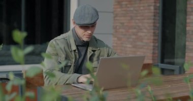 Laptopla çalışan serbest çalışan genç bir adam açık kafede oturuyor ve şehirdeki iş faaliyetlerine odaklanmış durumda. Serbest meslek ve modern teknoloji konsepti.