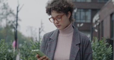 Şehir sokağında elinde kahve fincanıyla akıllı telefonuyla mesajlaşan genç bir iş kadını. İş iletişimi ve modern yaşam tarzı kavramı.